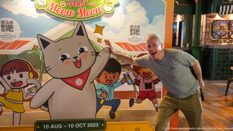 Gray-haired man playfully posing beside a large cartoon cat on a hawker-themed display.