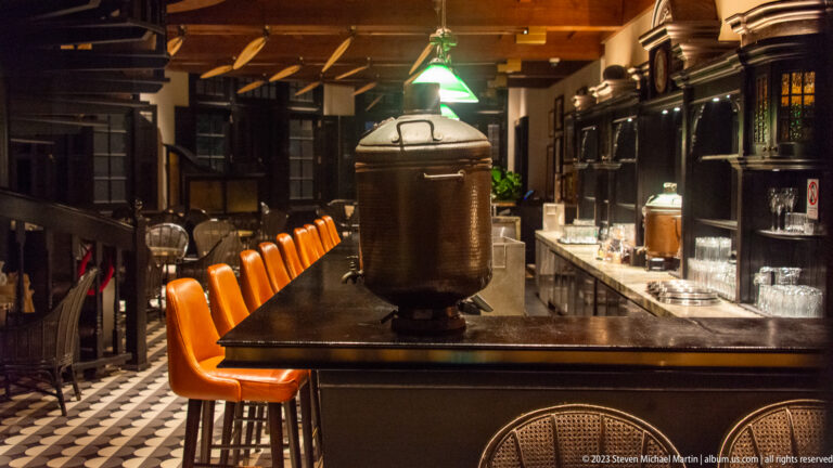 Dark wood Long Bar counter with orange stools, green lamps, glassware, and patterned floor.