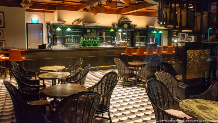 Long Bar seating area with marble tables, woven chairs, patterned floor, and dark wood counter.