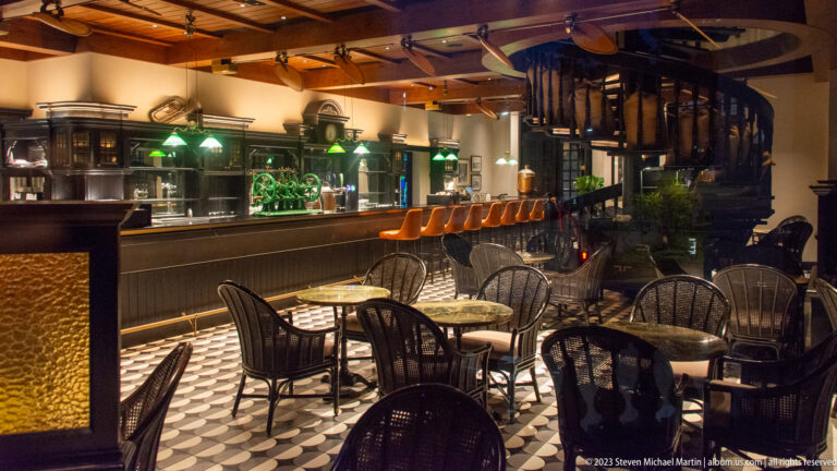 Wide Long Bar interior with tables, bar counter, orange stools, ceiling fans, and spiral staircase.