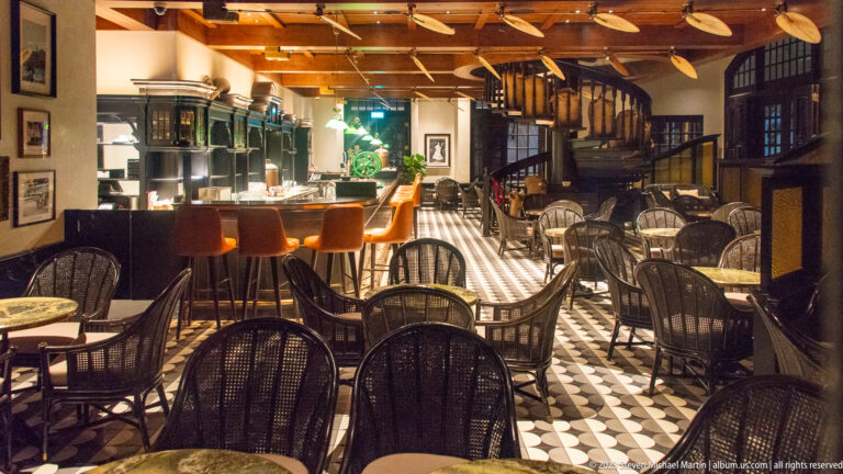 Wide Long Bar interior with wicker chairs, orange stools, patterned floor, ceiling fans, and spiral staircase.