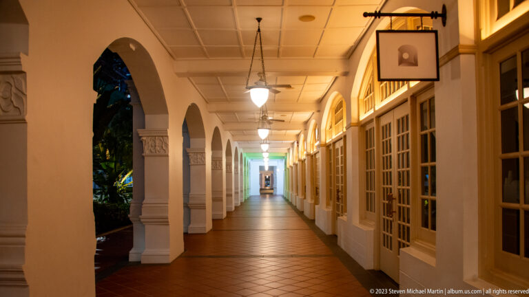 Long illuminated corridor with white arches, hanging lamps, and dark wood doors at Raffles Singapore.