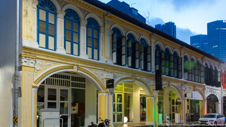 Illuminated two-story shophouse with arches, shuttered windows, pilasters, and ornate plaster decoration.
