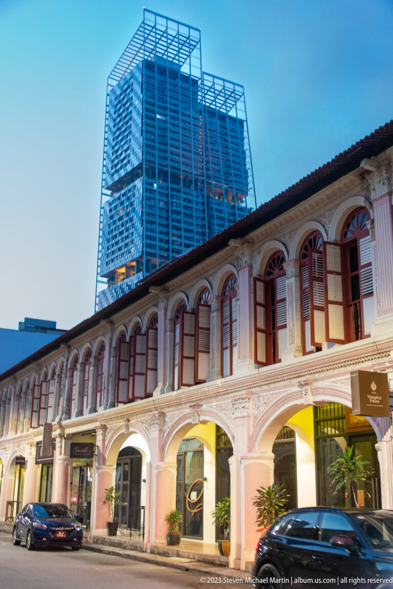Pastel shophouse row with arched bays and red windows, backed by a modern glass tower.