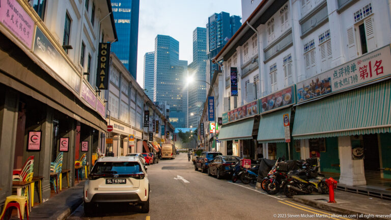 Narrow Singapore street with shophouses, awnings, parked motorcycles, cars, and skyscrapers beyond.