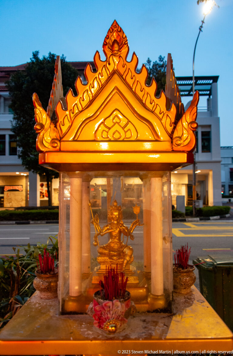 Small illuminated Chinese-style shrine with orange roof, incense sticks, and offerings.