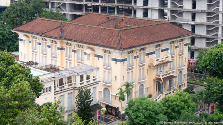 Historic cream-colored hotel with red tile roof, arched openings, balconies, and a construction site behind it.