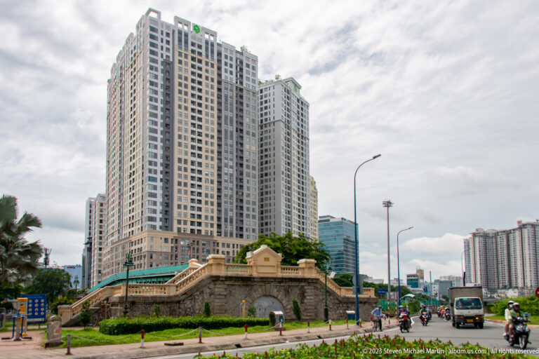 Low arched masonry structure beside traffic, backed by tall apartment buildings.