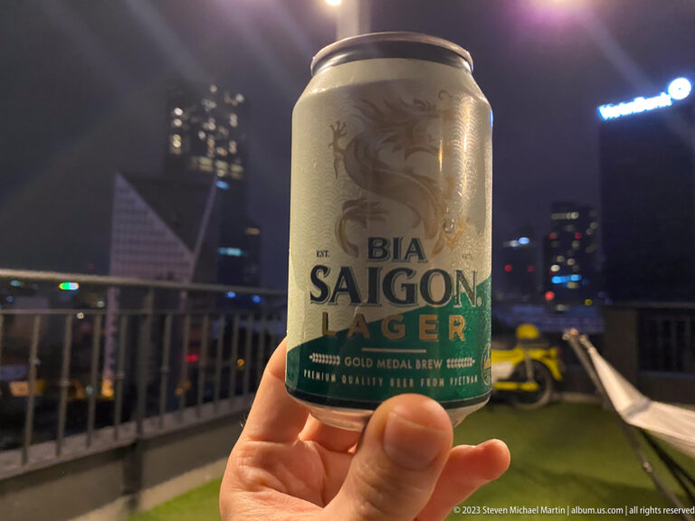 Hand holding a Bia Saigon Lager can against blurred rooftop lights at night.