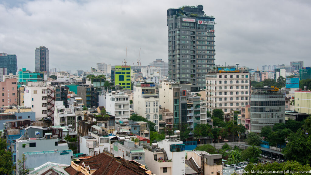 Dense Ho Chi Minh City skyline of low-rise neighborhoods and scattered modern towers under gray clouds.
