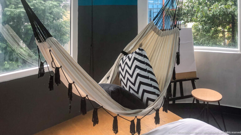 Cream tasseled hammock with patterned cushions beside a window and small table.