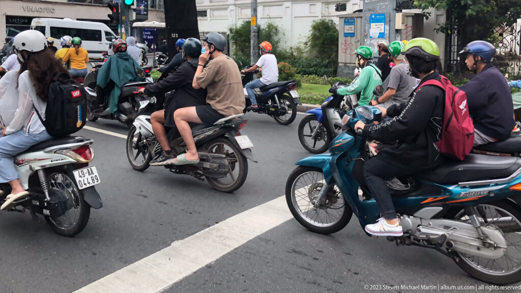Helmeted motorcyclists and passengers waiting closely together at a city intersection.