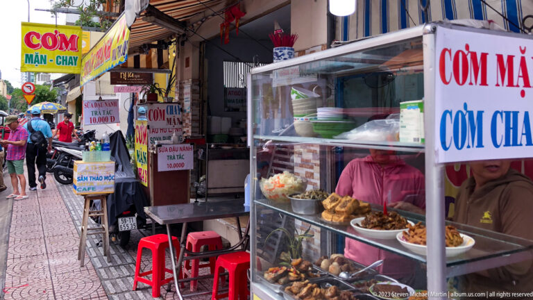 Sidewalk rice stall with glass food case and signs for meat and vegetarian meals.