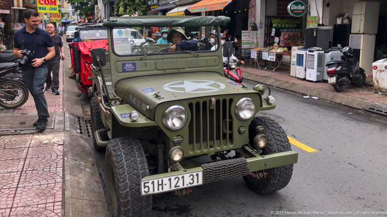 Olive-drab vintage jeep with white star driving past shops and parked motorbikes.