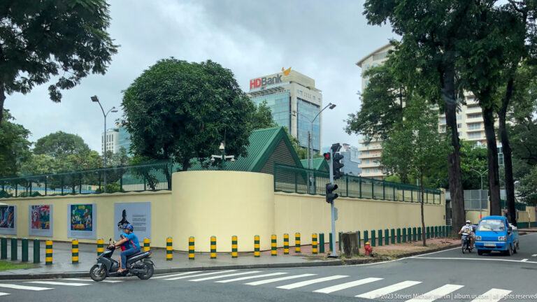 Cream walled compound with green-roofed guard pavilion, motorcyclist, trees, and office towers.