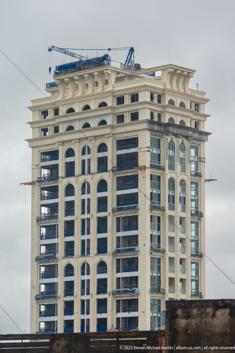 Unfinished cream-colored high-rise with arched façade openings, exposed floors, and construction equipment on the roof.