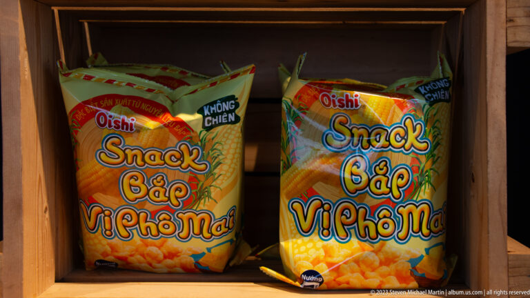 Two orange Oishi bags of cheese-flavored corn snacks on a wooden shelf.