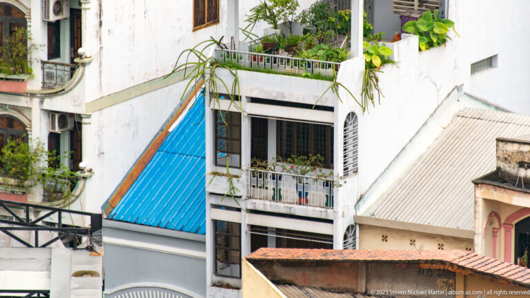 Bright blue corrugated awning among pale residential façades, balconies, shutters, and potted plants.
