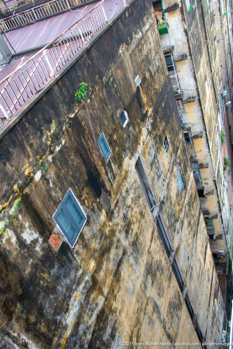 Angled view of a heavily weathered concrete wall with small windows, pipes, stains, and adjoining roofs.