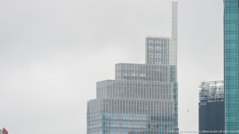 Stepped pale-glass high-rise with repeated façade grid and tall vertical rooftop element against a gray sky.