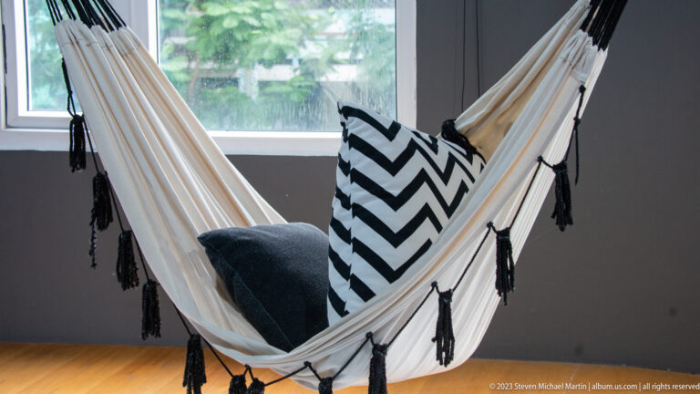 White indoor hammock with tassels and black-and-white chevron cushions positioned beside a large window.