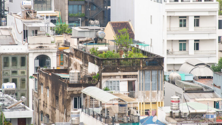 Dense cluster of weathered urban rooftops with water tanks, awnings, balconies, pipes, and potted plants.