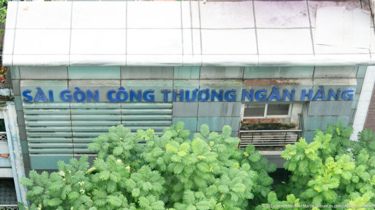 Blue Vietnamese bank lettering on a weathered louvered façade, partly obscured by leafy trees.
