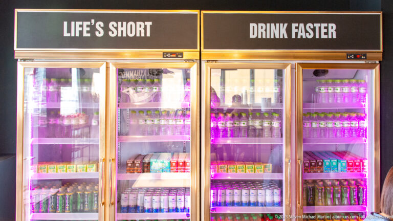 Purple-lit beverage refrigerators filled with drinks beneath signs reading “Life Is Short” and “Drink Faster.”
