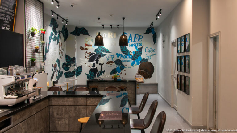 Narrow café with espresso counter, pendant lights, tables, framed pictures, and a coffee-themed wall mural.