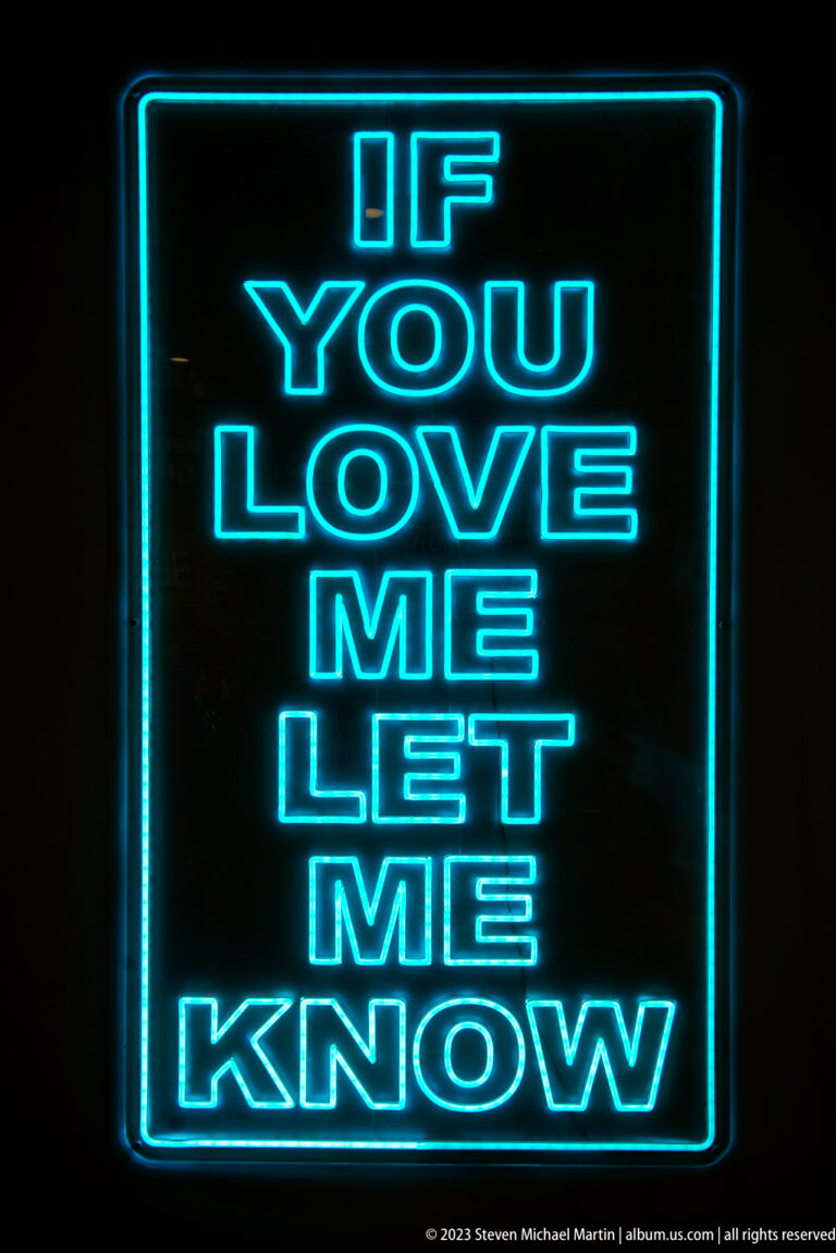 Blue neon sign on black reading “IF YOU LOVE ME LET ME KNOW” in stacked capital letters.