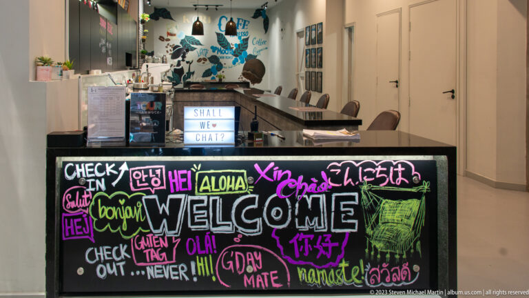 Black hotel reception counter covered with colorful multilingual greetings, with café equipment and corridor behind.