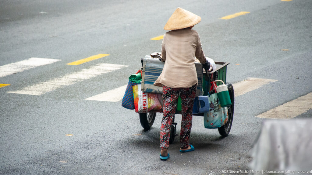Woman in a conical hat pushes a loaded two-wheeled cart across a gray city street.
