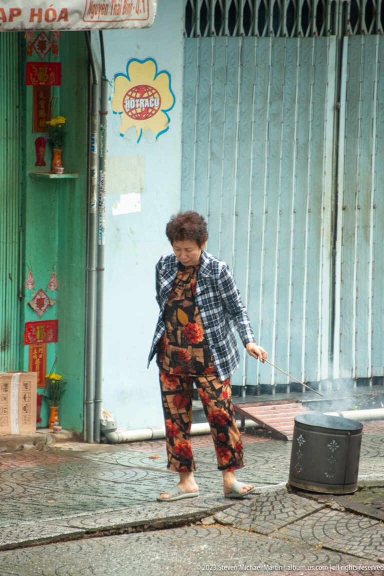 Older woman in patterned clothes gestures beside a portable speaker in front of a closed metal storefront.