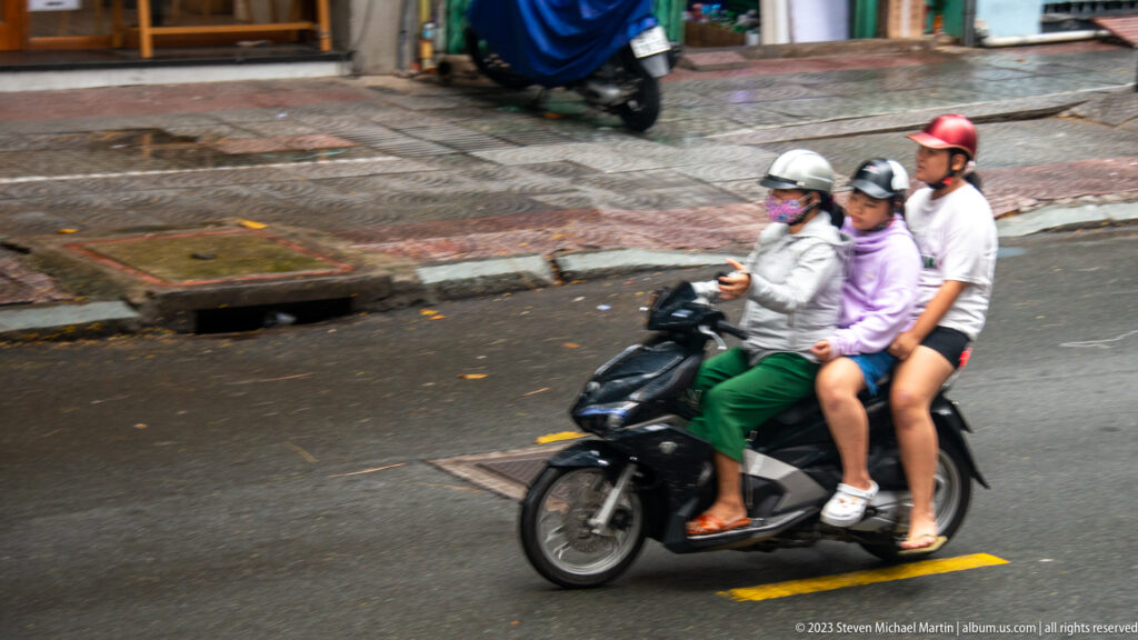 Three helmeted riders share a black motorbike on a city street viewed from above.