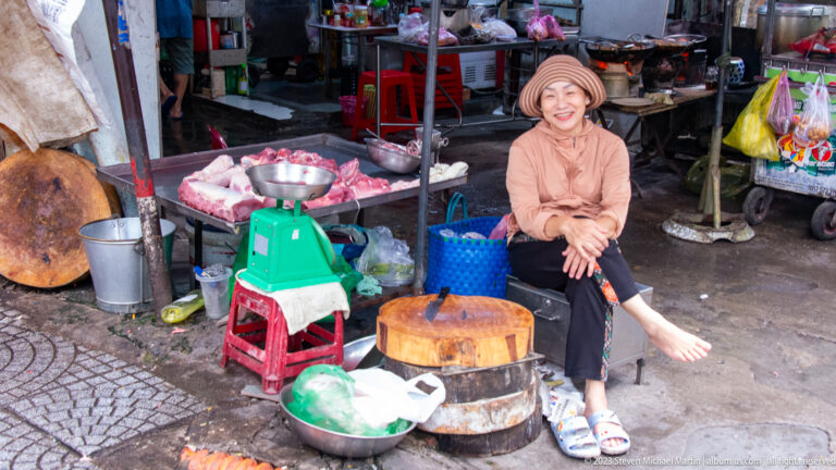 Smiling seated meat vendor beside chopping blocks, metal bowls, a green scale, baskets, and raw meat.