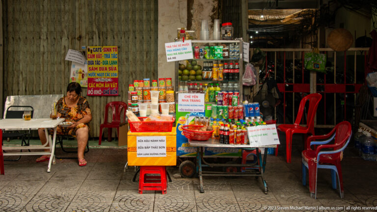 Small sidewalk shop with bottled drinks, packaged foods, Vietnamese notices, red stools, and a seated woman.