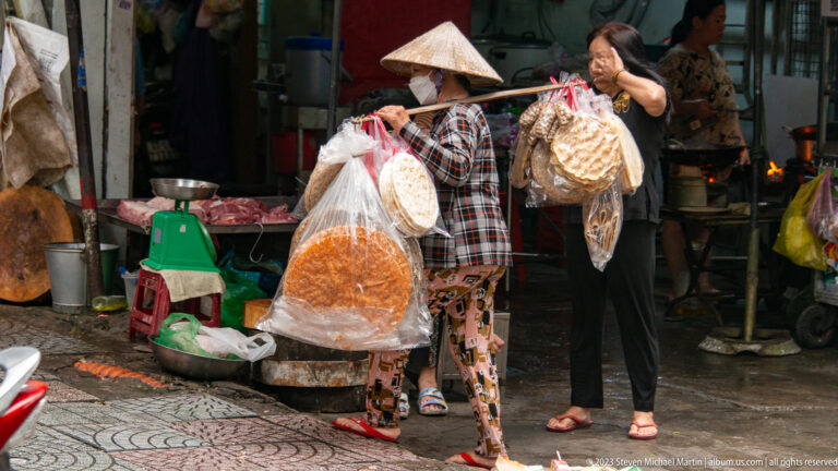 Two women handle large bags of round dried foods beside chopping blocks, bowls, and a market scale.