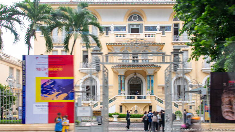 Cream-and-yellow historic building with arches, columns, balconies, balustrades, and red roof behind trees and a display panel.