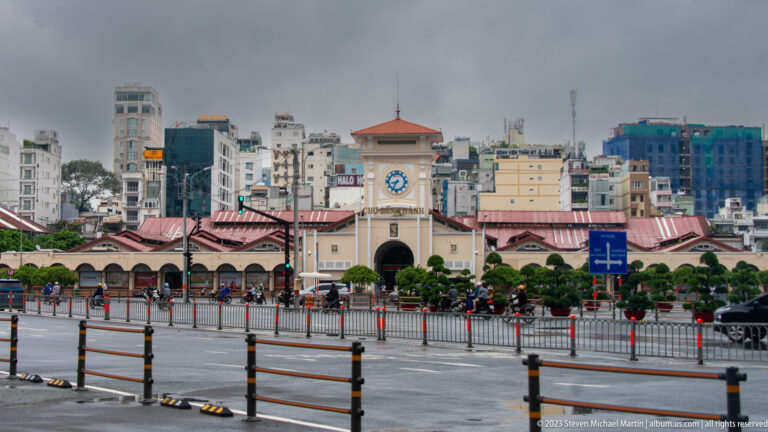 Bến Thành Market south façade with central clock tower, red roofs, arched entrance, and city buildings behind.