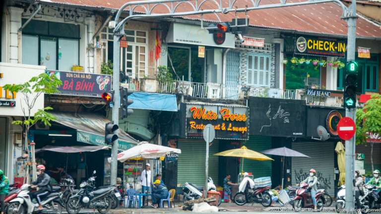 Busy row of narrow shops and restaurants with signs, balconies, awnings, parked motorbikes, and street seating.