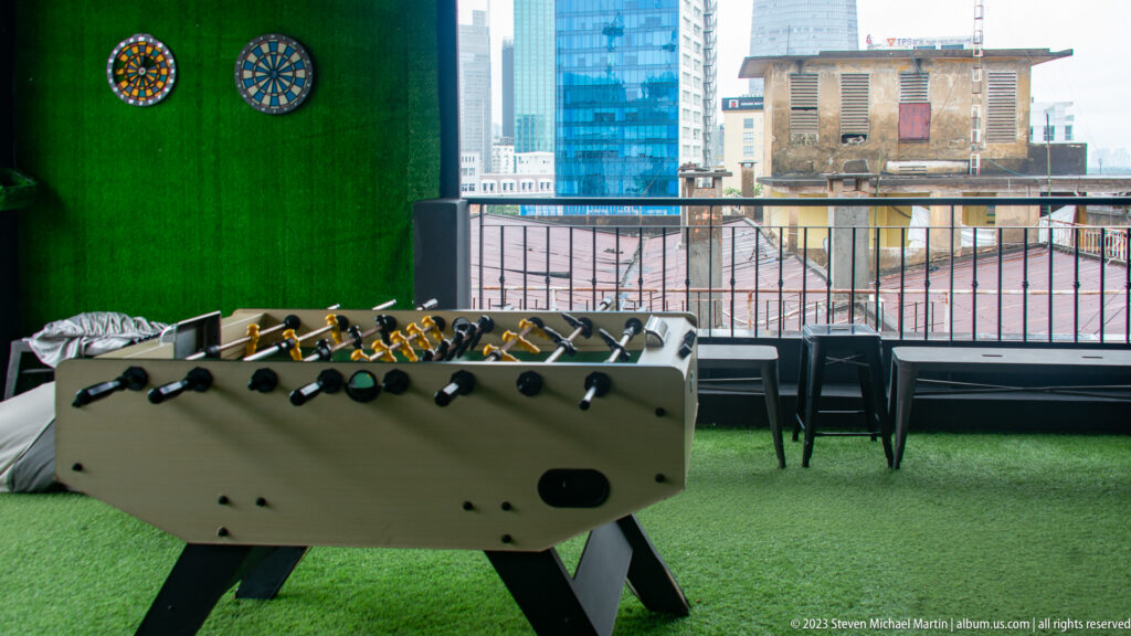 Foosball table on green artificial turf beside a rooftop railing, with dense city buildings in the background.