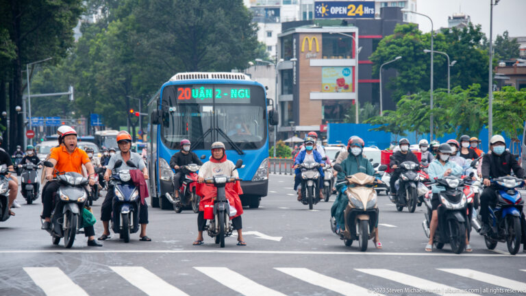 Large group of helmeted motorbike riders waits at a zebra crossing with a city bus behind.