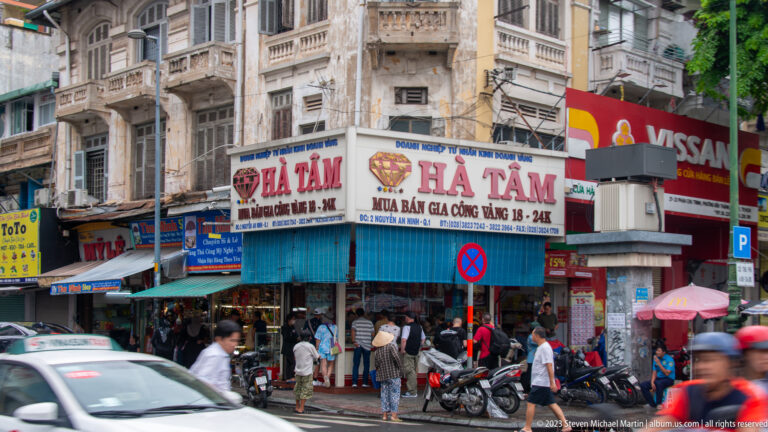 Busy Hà Tâm gold and currency exchange storefront with customers, blue awnings, motorbikes, and upper-floor balconies.