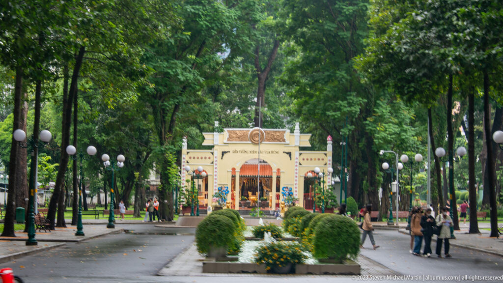 Yellow-and-cream park gateway at the end of a tree-lined avenue with globe lamps, shrubs, flowers, and pedestrians.