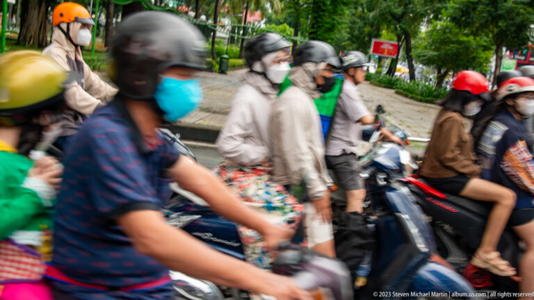 Close blurred view of helmeted and masked motorbike riders passing across a green urban streetscape.