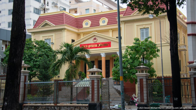 Yellow institutional building with red-tiled roof, classical portico, columns, fence, trees, and red Vietnamese sign.