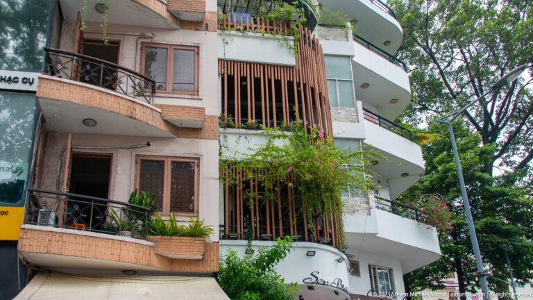 Curved residential balconies with vertical screens, brick walls, potted plants, vines, and large street trees.