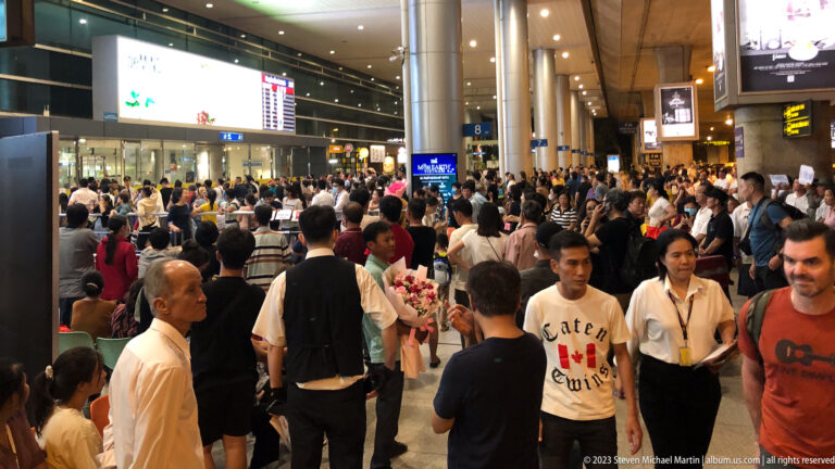 Crowded airport arrivals hall with waiting families, passengers, flowers, signs, columns, and terminal lighting.