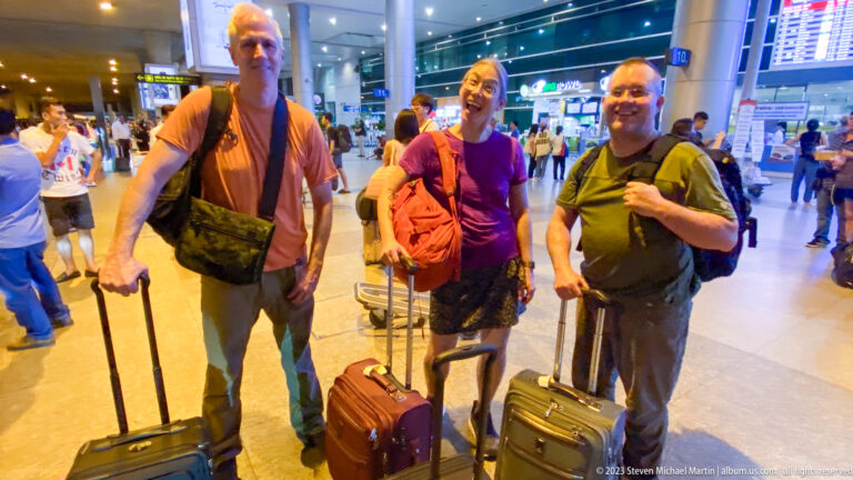 Three smiling male travelers stand with rolling suitcases and shoulder bags inside a busy airport terminal.
