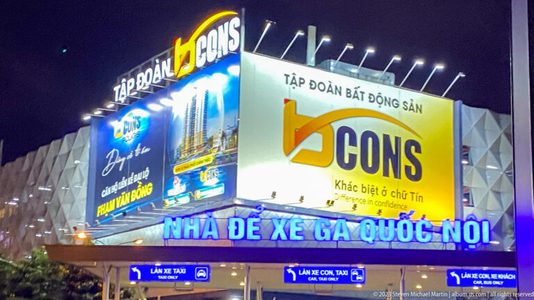 Nighttime domestic-terminal parking entrance with bright Vietnamese signs, blue vehicle-lane directions, and large illuminated advertisements.